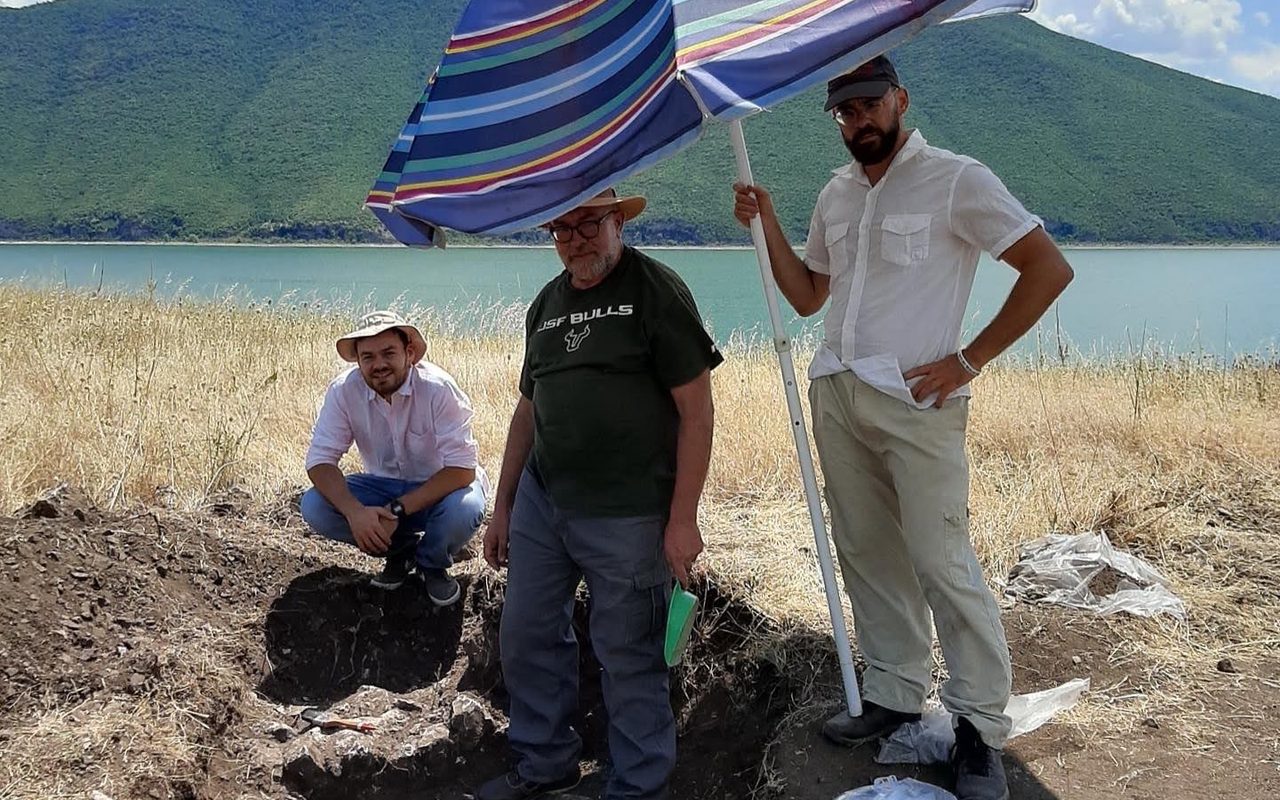 Three archaeologists at the trench on Maligrad under a striped beach umbrella, Lake Prespa behind them